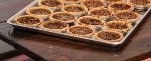 A tray of small, or "penny", pecan pie tins sits on a wooden picnic table.