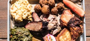 A silver tray filled with mac and cheese, hushpuppies, toast, greens, pork, brisket, chicken, veggies and more.