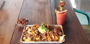 Mount Nacho - featuring homemade tortilla chips, queso, corn salsa, pickled guacamole, jalapeño and Pulled Pork - sits on a silver tray near a bowl of pecans and a Bloody Mary.