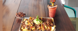 Mount Nacho - featuring homemade tortilla chips, queso, corn salsa, pickled guacamole, jalapeño and Pulled Pork - sits on a silver tray near a bowl of pecans and a Bloody Mary.