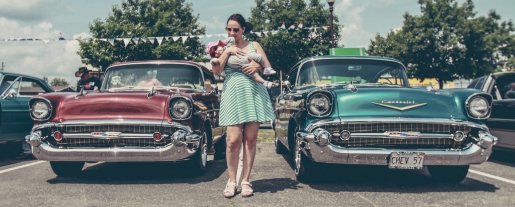 A woman in a teal romper carries her child between two vintage, '57 Chevys.
